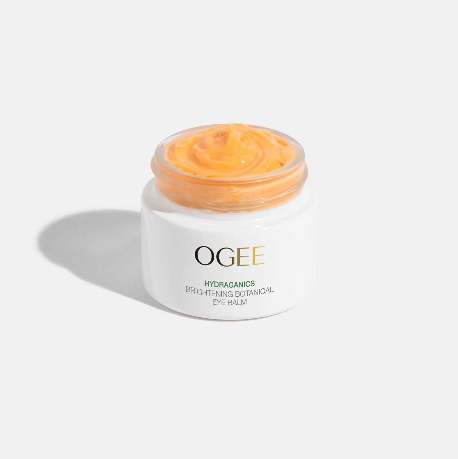 A jar of OGEE brand Brightening Botanical Eye Balm with a golden applicator, the product is visible as a creamy orange color.